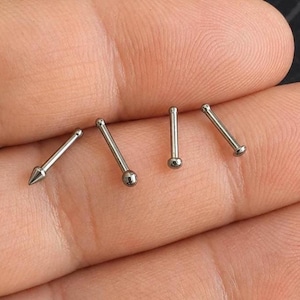 May include: A selection of silver nose studs in different styles. The set features a pointed stud, a stud with a small ball, and two studs with a tiny ball. The jewellery is shown on a hand.
