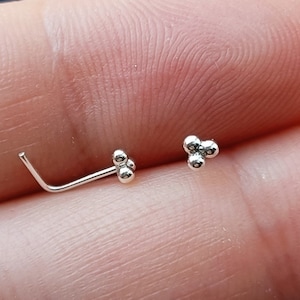 May include: Two silver nose rings. One is a simple L-shaped ring with a small ball at the end. The other is a cluster of three small balls.