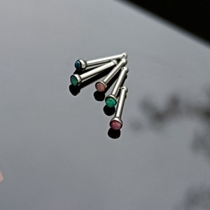 May include: A collection of silver nose studs with colorful, round gemstones. The gemstones are in shades of blue, green, pink, and red. The studs are arranged on a reflective black surface.