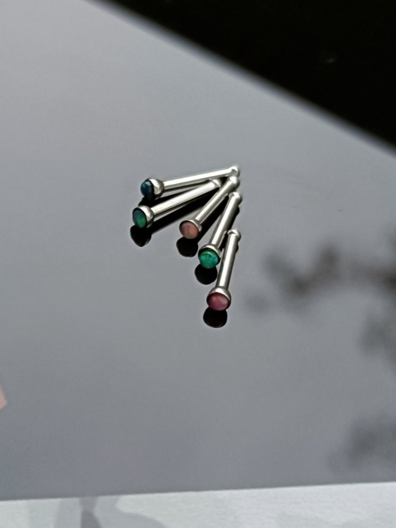 May include: A collection of silver nose studs with colorful, round gemstones. The gemstones are in shades of blue, green, pink, and red. The studs are arranged on a reflective black surface.