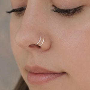 May include: Close-up of a nose with two silver nose rings. The rings are small and circular, positioned close together on the nostril. The skin is fair, and the background is a soft, neutral tone. The image focuses on the jewelry.
