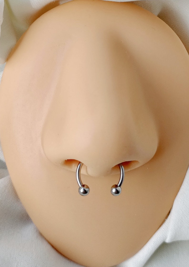 Pu&ograve; includere: Primo piano di un piercing al naso con due anelli d'argento. Gli anelli sono circolari con piccole sfere ad ogni estremit&agrave;.
