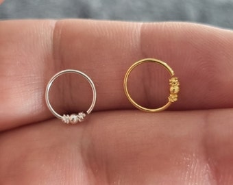 May include: Two nose rings, one silver and one gold, with a small bead accent. The rings are shown on a finger.