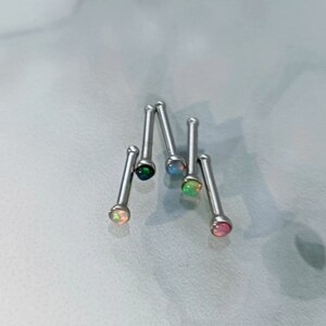 May include: A collection of nose studs with colorful opal gemstones. The studs have a silver-colored post and a round, iridescent opal stone. The opals display a range of colors, including pink, green, blue, and black. The studs are arranged on a light-colored surface.