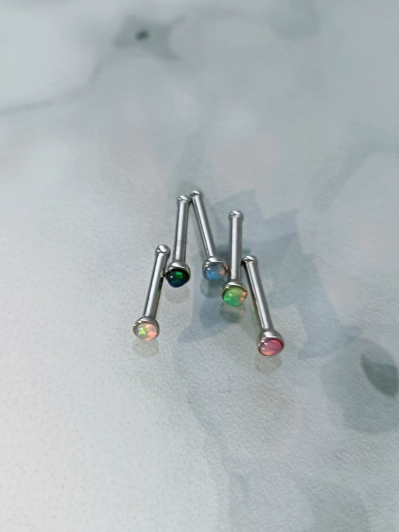 May include: A collection of nose studs with colorful opal gemstones. The studs have a silver-colored post and a round, iridescent opal stone. The opals display a range of colors, including pink, green, blue, and black. The studs are arranged on a light-colored surface.