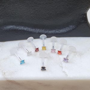 May include: A selection of lip piercing retainers featuring different coloured square-cut gemstones. The retainers have clear, flexible posts and flat, round discs. Gemstone colours include blue, silver, red, yellow, pink, orange, and purple.