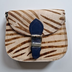 Puede incluir: Un pequeño bolso bandolera de cuero con estampado de cebra marrón y blanco con cierre de hebilla de latón azul marino.