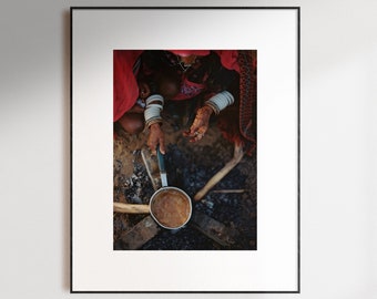 May include: A person wearing traditional clothing and jewelry stirs a pot over a fire. The pot is made of metal and has a dark brown interior. The fire is made of wood and is burning brightly.