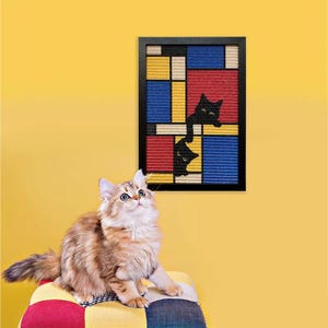 以下が含まれることがあります： パッチワークのクッションに座り、上を見上げるふわふわのマルチカラーの猫。赤、青、黄、黒のモンドリアン風デザインで、黒猫のシルエットが描かれた額入りのアートワークが、黄色の壁に掛けられています。