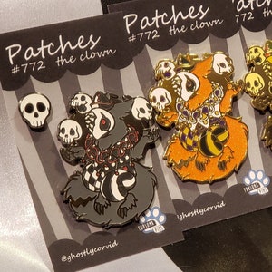 May include: Four enamel pins featuring a cartoon character named Patches the clown. The character is a furry creature with a skull-themed design. The pins are in different colors: black, orange, brown, and purple. The pins are on a black and white striped background.