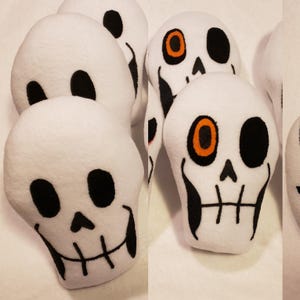 May include: Five white plush skull-shaped pillows with black eyes and mouths. One skull has an orange eye with a black center.