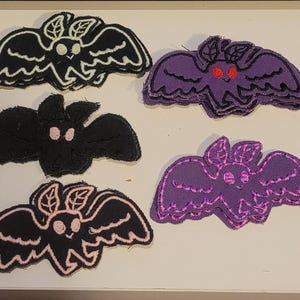 Puede incluir: Una colección de apliques de fieltro hechos a mano con forma de murciélago en varios colores. Los murciélagos son negros, morados y blancos, algunos con detalles en rosa o rojo. Los apliques tienen diseños detallados de alas y caras.