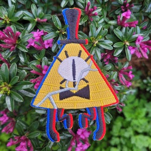Könnte beinhalten: Ein gelbes Dreieck mit einem einzigen Auge und einer schwarzen Fliege. Das Dreieck hat einen schwarzen Zylinder mit roten und blauen Verzierungen. Das Dreieck ist auf einem Stoffpatch gestickt.