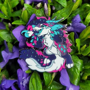 May include: A colourful enamel pin featuring a cartoon animal with blue, pink and purple fur. The animal has a white face with black eyes and a pink tongue sticking out. The pin is set against a background of purple flowers.