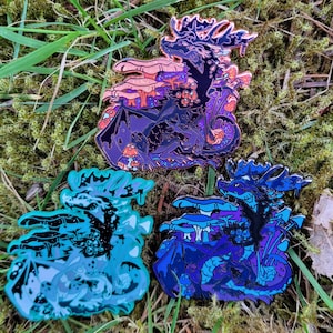 Puede incluir: Tres pines de esmalte con un dragón negro con detalles en morado y azul rodeado de setas. El dragón está en un entorno de bosque. Los pines tienen un diámetro de aproximadamente 2,5 cm.