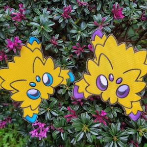 Peut inclure: Deux patchs brodés jaunes et violets représentant un personnage de dessin animé avec trois yeux. Le personnage a un aspect duveteux et hérissé.