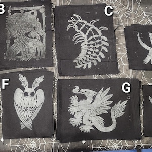 May include: A selection of black fabric squares featuring white graphic prints. Designs include a rat, a fish, a crustacean, a rat with the word "RAT!", another rat, an owl, a griffin, and a rat with the word "AAAAAA".