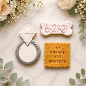 May include: Three decorated biscuits: one shaped like a diamond ring with white and grey icing, one bone-shaped biscuit with white icing and the date 6-27-21 in pink, and one square biscuit with scalloped edges featuring the text MY HUMANS ARE ENGAGED.