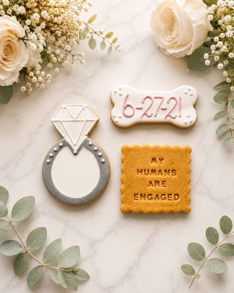 May include: Three decorated biscuits: one shaped like a diamond ring with white and grey icing, one bone-shaped biscuit with white icing and the date 6-27-21 in pink, and one square biscuit with scalloped edges featuring the text MY HUMANS ARE ENGAGED.