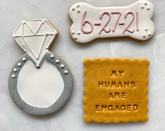 May include: Three decorated cookies. One is a bone-shaped cookie with the date "6-27-21" in pink icing. Another is a round cookie with a gray ring and a white diamond in the center. The last cookie is a square cookie with the text "My humans are engaged" in pink icing.