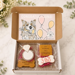 May include: A cardboard gift box containing two clear plastic bags of dog treats. One bag holds a diamond ring-shaped biscuit and a bone-shaped biscuit labelled MUST LOVE DOGS. The other holds a square biscuit reading MY HUMANS ARE ENGAGED and a bone-shaped biscuit with 3-18-23. The box lid features a dog illustration and the text CONGRATULATIONS.