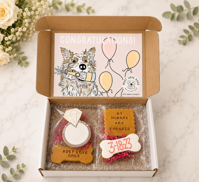 May include: A cardboard gift box containing two clear plastic bags of dog treats. One bag holds a diamond ring-shaped biscuit and a bone-shaped biscuit labelled MUST LOVE DOGS. The other holds a square biscuit reading MY HUMANS ARE ENGAGED and a bone-shaped biscuit with 3-18-23. The box lid features a dog illustration and the text CONGRATULATIONS.