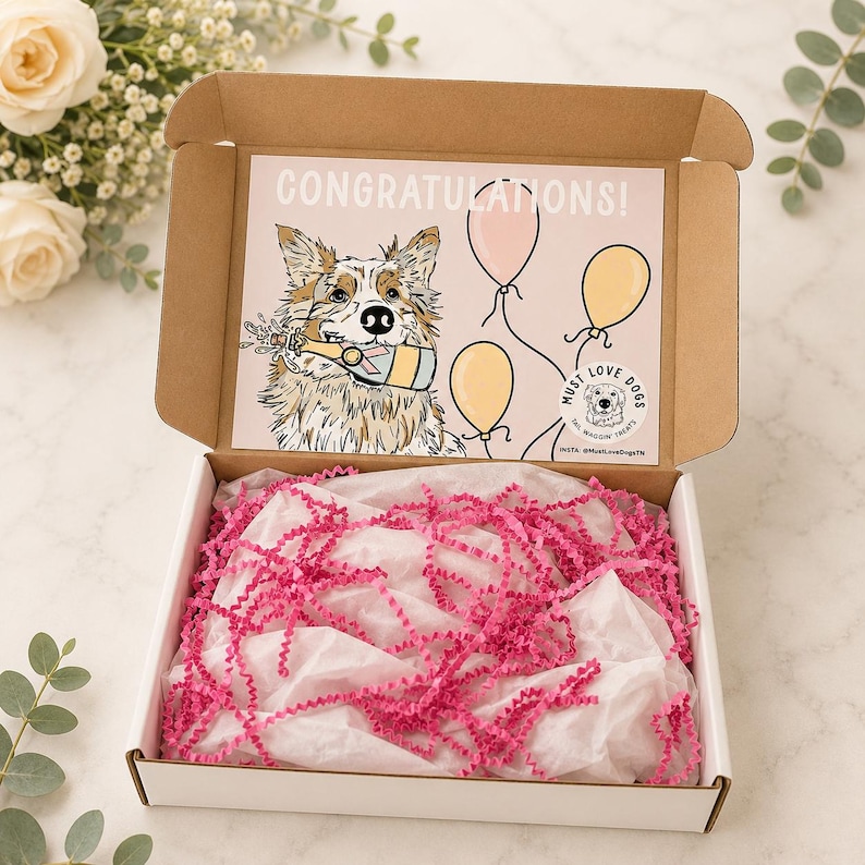 May include: A cardboard gift box with a white interior and brown exterior. The lid features a printed illustration of a dog holding a champagne bottle with the text CONGRATULATIONS and three balloons. The box contains white tissue paper and pink crinkle paper.