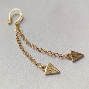 May include: Gold-colored ear cuff with two delicate chains, each ending in a textured arrow charm. The ear cuff features a curved design for secure wear. The chains hang down, creating a stylish, minimalist look.