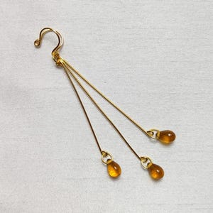 Puede incluir: Pendientes colgantes dorados con tres finas hebras, cada una terminada en una cuenta ámbar en forma de lágrima. Los pendientes tienen un cierre de gancho y están sobre un fondo blanco.