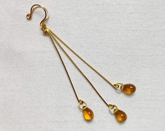May include: Gold-colored dangle earrings with three delicate strands, each ending in a teardrop-shaped amber bead. The earrings have a hook closure and are set against a white background.