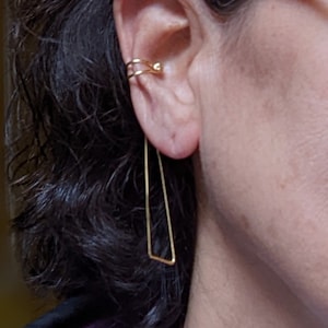 May include: Gold ear cuff and a long, thin, gold, rectangular earring. The earring is hanging from the earlobe.