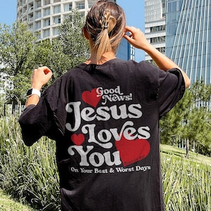 May include: Black t-shirt with a white graphic that says "Good News! Jesus Loves You" with red hearts. The text below the graphic says "On Your Best & Worst Days".