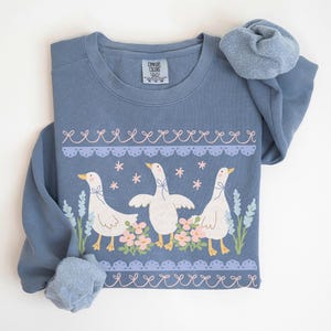 May include: A blue long-sleeved sweatshirt featuring a whimsical design of three white geese with floral and lace accents. The geese are standing among pink flowers and blue sprigs. The sweatshirt has a crew neck and a relaxed fit.