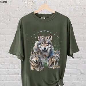 May include: A green t-shirt with a graphic print of three wolves in a forest setting. The wolves are surrounded by trees and a crescent moon. The shirt is made of a soft, comfortable material and is perfect for everyday wear.