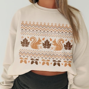May include: A cream-colored sweatshirt with a brown and orange autumn-themed design featuring squirrels, acorns, and leaves. The design is a repeating pattern of squirrels, acorns, and leaves.
