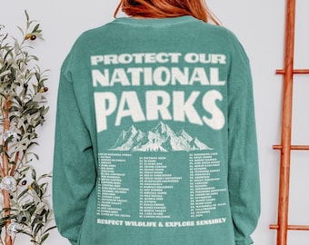 May include: A teal long-sleeve shirt with the text "PROTECT OUR NATIONAL PARKS" and a mountain graphic. Below, a list of national parks is printed. The phrase "RESPECT WILDLIFE & EXPLORE SENSIBLY" is also included.