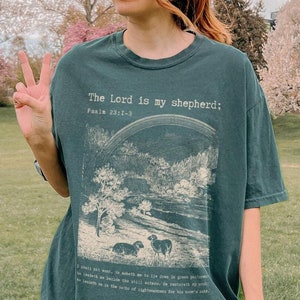 Puede incluir: Una camiseta verde oscuro con una ilustraci&oacute;n vintage de un pastor y ovejas. El texto "The Lord is my shepherd; Psalm 23:1-3" est&aacute; impreso en la parte delantera de la camiseta.