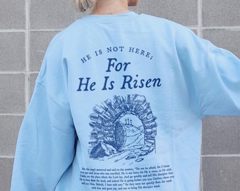 Pode incluir: Camisola azul claro com o texto "He is not here, For He is Risen" e um desenho de um túmulo de pedra com uma porta aberta. O texto "Matthew 28:6" também está incluído.