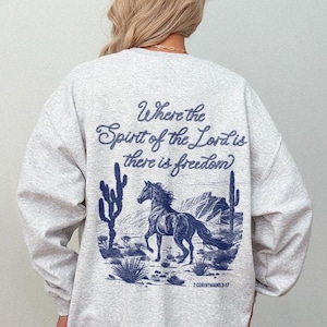 May include: Gray sweatshirt with a blue graphic print of a horse running through a desert landscape. The text "Where the Spirit of the Lord is there is freedom" is printed on the back of the sweatshirt.