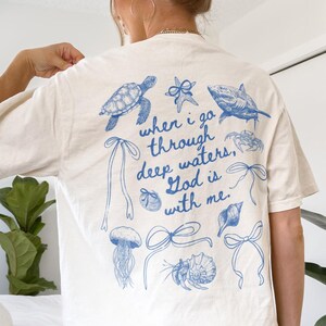 May include: Cream-colored t-shirt with blue illustrations of sea creatures and bows. The text "when I go through deep waters, God is with me" is printed in blue. The shirt is a casual, short-sleeved style.