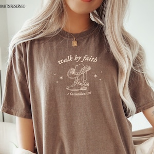 Pu&ograve; includere: Una maglietta marrone con una grafica di stivali da cowboy e un cappello con il testo "walk by faith" e "2 Corinthians 5:7".