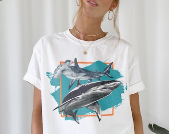May include: White t-shirt with a graphic of two sharks, one hammerhead and one with a pointed nose, in a turquoise and orange frame. The graphic is on a white t-shirt with a crew neck.