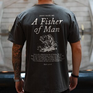 May include: A dark grey t-shirt with a white graphic print of a fish jumping out of the water. The text "JESUS MADE ME A FISHER OF MAN" is printed above the fish. The text "Mark 1:16-18" is printed below the fish.