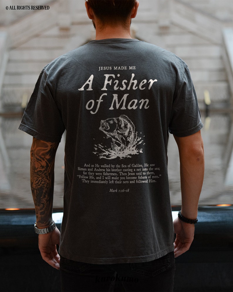 May include: A dark grey t-shirt with a white graphic print of a fish jumping out of the water. The text "JESUS MADE ME A FISHER OF MAN" is printed above the fish. The text "Mark 1:16-18" is printed below the fish.