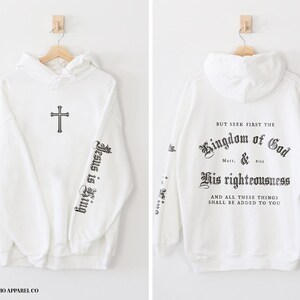Op de afbeelding: Witte hoodie met een kruis op de voorkant en de tekst "Jesus is King" op de linkermouw. De achterkant van de hoodie heeft de tekst "But seek first the Kingdom of God & His righteousness and all these things shall be added to you".