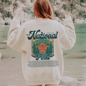 Może przedstawiać: Kremowa bluza z nadrukiem niedźwiedzia w leśnej scenerii oraz napisem "U.S. National Parks Since 1972". Bluza wymienia r&oacute;żne parki narodowe. Marka "kurokumo apparel co" jest wydrukowana na dole.