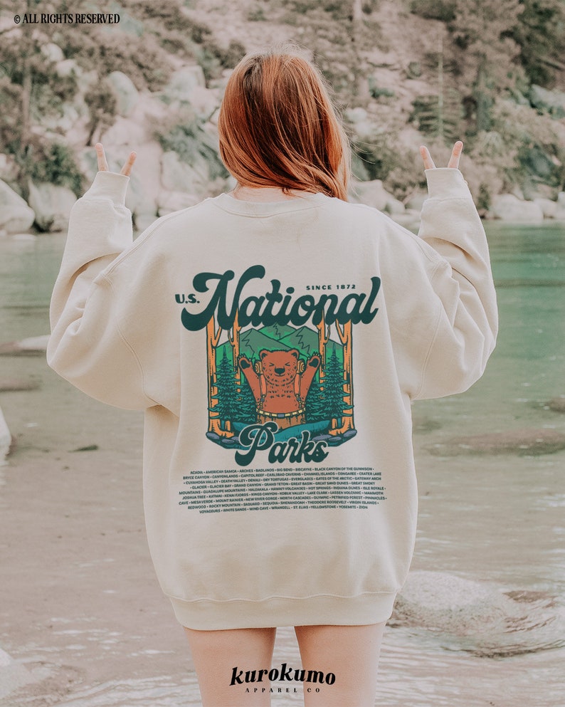 Może przedstawiać: Kremowa bluza z nadrukiem niedźwiedzia w leśnej scenerii oraz napisem "U.S. National Parks Since 1972". Bluza wymienia r&oacute;żne parki narodowe. Marka "kurokumo apparel co" jest wydrukowana na dole.