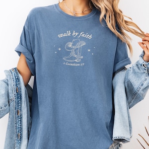 Pu&ograve; includere: Una maglietta blu chiaro con una grafica bianca di uno stivale da cowboy e un cappello con il testo "walk by faith" e "2 Corinthians 5:7".