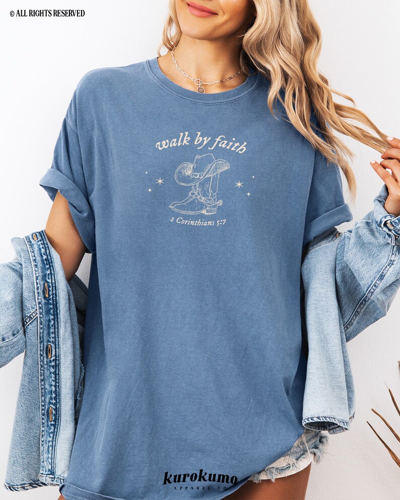 Pu&ograve; includere: Una maglietta blu chiaro con una grafica bianca di uno stivale da cowboy e un cappello con il testo "walk by faith" e "2 Corinthians 5:7".