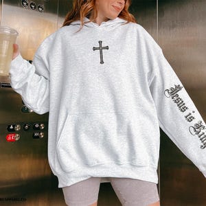 Op de afbeelding: Een grijze hoodie met een zwart kruis op de voorkant en de tekst "Jesus is King" op de mouw.